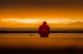 Sad man in red coat sits beside a lake at sunset.