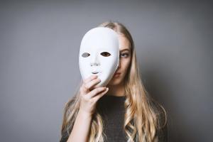 Teen blonde girl hiding her face behind a mask with a gray background