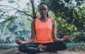 black woman in orange shirt and black leggings meditating outside in wooded area