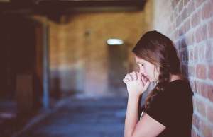 woman with dark braid in black shirt holding hands by face in a brick alleyway