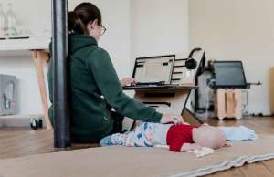 woman with dark hair in green sweatshirt sitting on laptop on floor touching baby