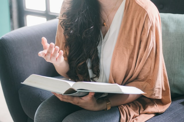 woman with black hair sitting on a grey couch and holding a book.