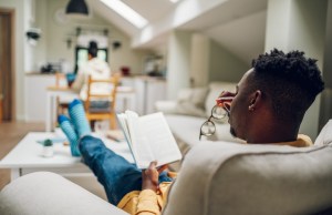 Back view of an African American man sitting in an armchair and reading a book at home while his wife is sitting in the background and using a laptop